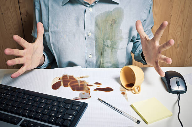 Person sits with coffee-stained shirt and spills on desk, illustrating need for household stain removal tips.